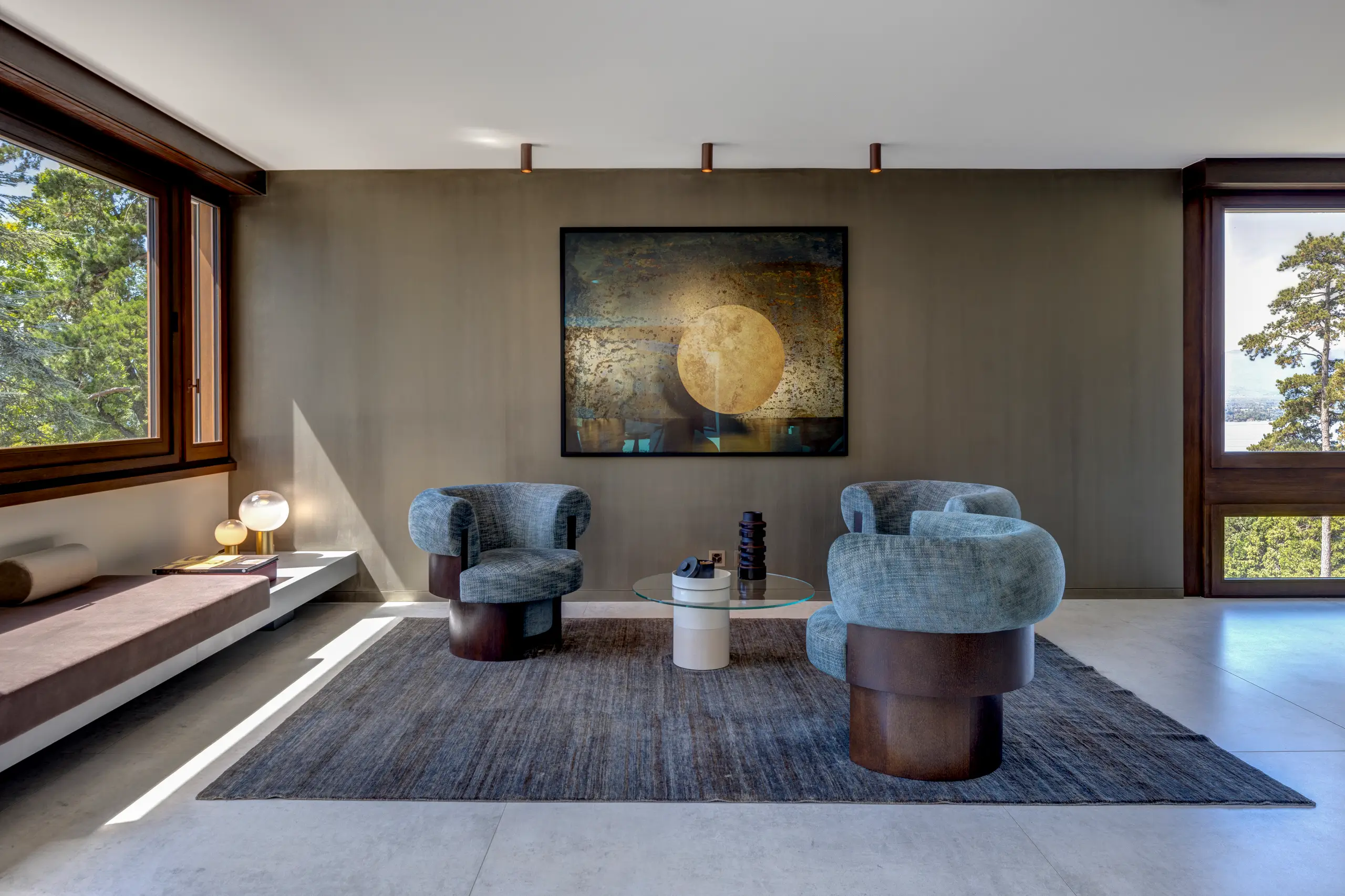 Sitting area with two grey lounge chairs and dark rounded side tables on a textured rug beside a timber-framed window